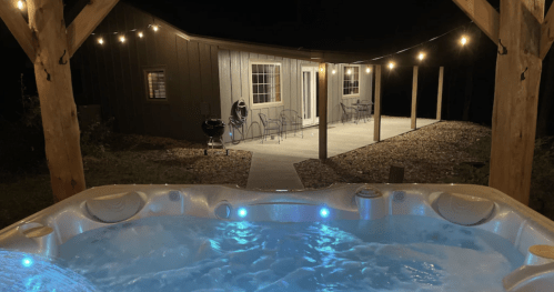 A hot tub with blue lights in the foreground, overlooking a cozy cabin with string lights at night.