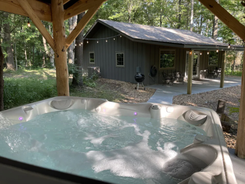 A hot tub with bubbling water in a wooded area, overlooking a cozy cabin with string lights and a grill.