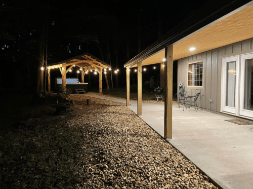 A well-lit outdoor area at night featuring a patio, gravel path, and a gazebo with string lights in a wooded setting.