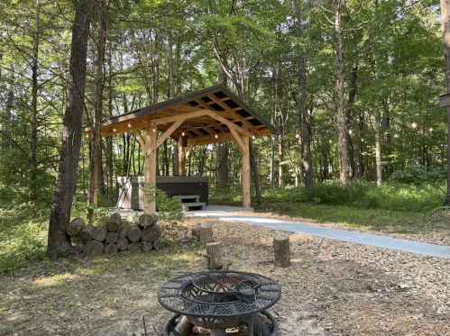 A wooden pavilion in a forested area, with a fire pit and stacked logs nearby, surrounded by greenery.