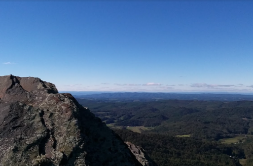 A rocky mountain peak overlooks a vast landscape of rolling hills and a clear blue sky.