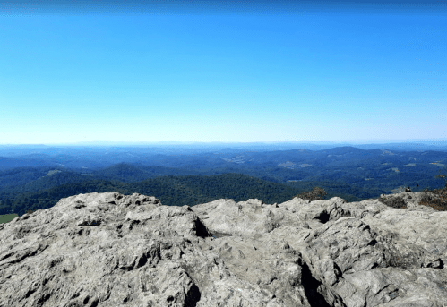 A panoramic view from a rocky summit, showcasing rolling hills and a clear blue sky.