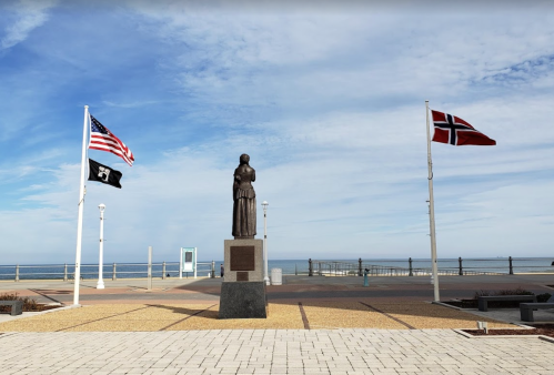 A statue stands between the American and Norwegian flags, overlooking a beach and ocean under a cloudy sky.