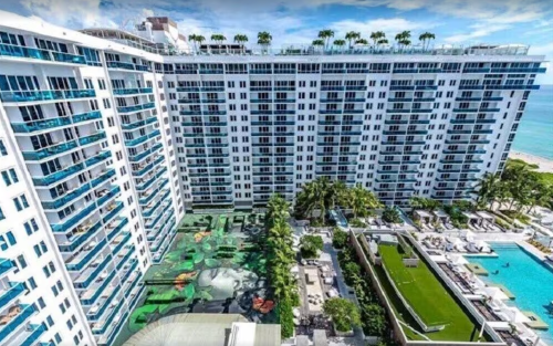 Aerial view of a modern hotel with balconies, palm trees, and a pool area near the beach.