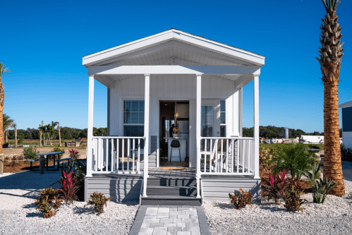 A small, modern white house with a porch, surrounded by palm trees and landscaped gravel. Clear blue sky above.