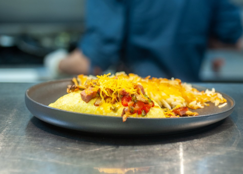 A plate of omelette topped with cheese, diced tomatoes, and crispy toppings, served with a side of hash browns.