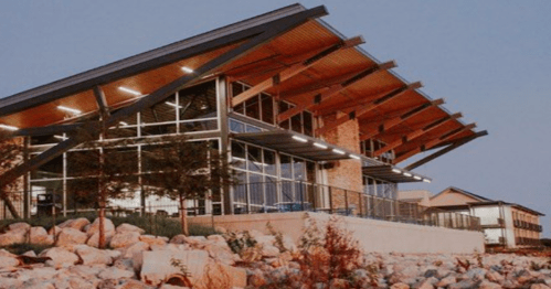 Modern building with a sloped roof, large windows, and a stone foundation, set against a twilight sky.