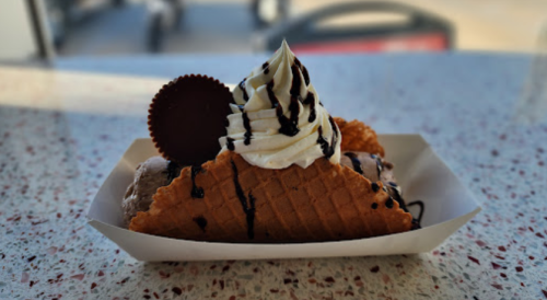 A waffle cone filled with chocolate ice cream, topped with whipped cream, chocolate drizzle, and a chocolate cup.