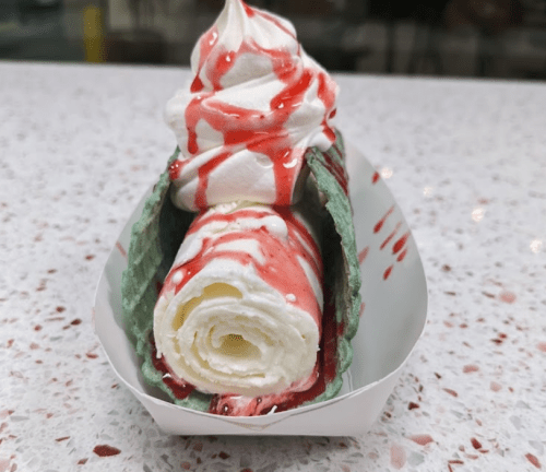 A rolled ice cream dessert in a green waffle cone, topped with whipped cream and drizzled with strawberry sauce.