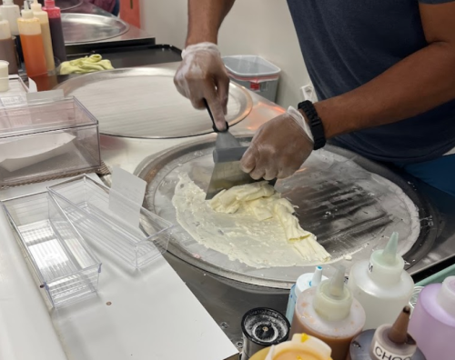 A person in gloves uses a spatula to spread ice cream on a cold metal surface in a dessert shop.