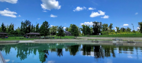 A serene lake reflecting blue skies and fluffy clouds, surrounded by lush greenery and trees.