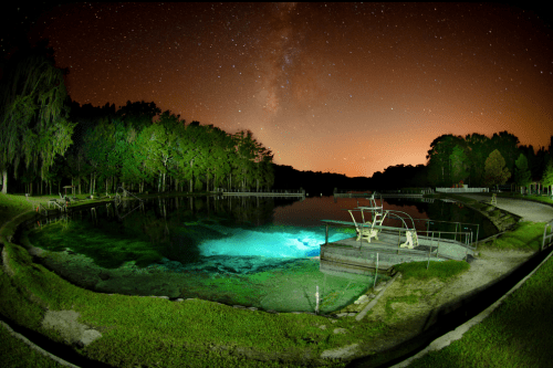 A serene lake at night, illuminated by the Milky Way and soft lights, surrounded by trees and a diving platform.