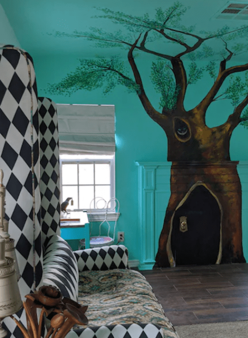 A cozy room with a turquoise wall featuring a large tree mural, a patterned chair, and a small table by the window.