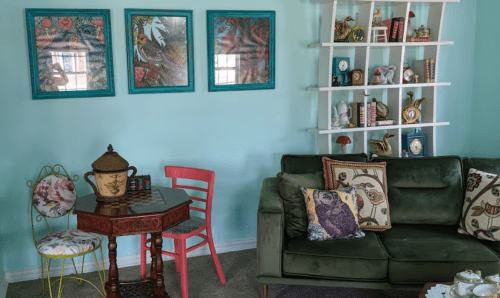 A cozy living room with teal walls, a green sofa, decorative shelves, and a small table with colorful chairs.