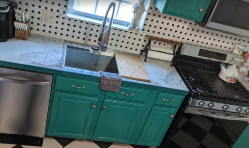 A modern kitchen with teal cabinets, a stainless steel sink, and a marble countertop, featuring a gas stove and black-and-white tiles.
