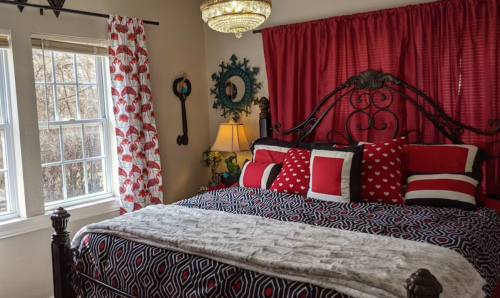 Cozy bedroom featuring a black and red bedspread, decorative pillows, a chandelier, and large windows with curtains.