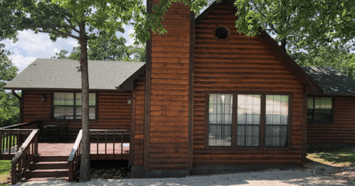 A rustic log cabin with a wooden deck, surrounded by trees and a clear blue sky.