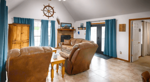 Cozy living room with brown leather chairs, blue curtains, a wooden cabinet, and nautical decor. Bright and inviting space.