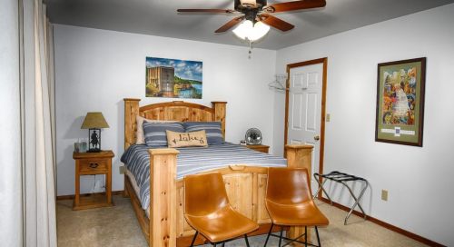 Cozy bedroom with a wooden bed, two chairs, a nightstand, and wall art, featuring a ceiling fan and neutral decor.