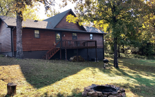 A wooden cabin with a deck, surrounded by trees and a grassy area, featuring a fire pit in the foreground.