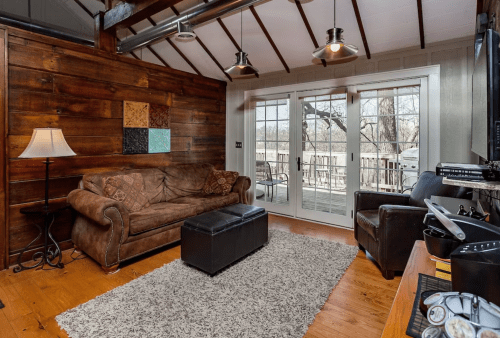 Cozy living room with a brown sofa, ottoman, and large windows overlooking a deck, featuring wooden walls and a rug.