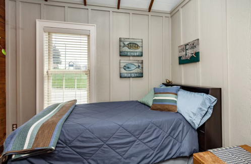 Cozy bedroom with a blue bedspread, striped pillows, and fish-themed wall art. Natural light from a window.