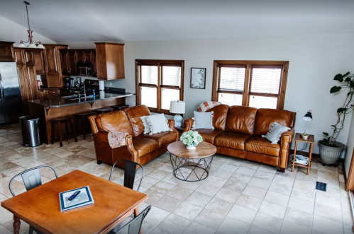 Cozy living room with brown leather sofas, a round coffee table, and a kitchen area in the background. Bright and inviting.