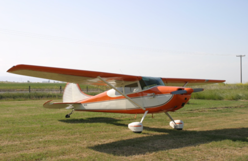 A small orange and white airplane parked on a grassy field under a clear sky.
