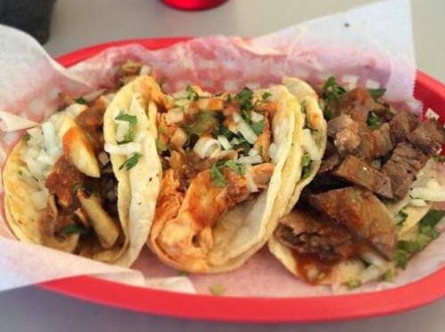 A red basket filled with three tacos topped with onions and cilantro, featuring different meats and sauces.