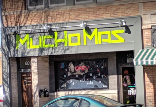 Sign for "Mucho Mas" with bright green letters on a brick building, featuring an "Open" sign in the window.