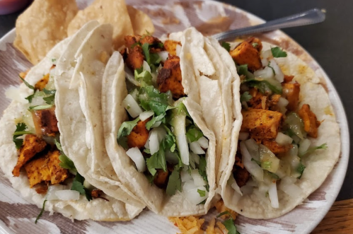 Three tacos filled with seasoned meat, onions, and cilantro, served on a plate with tortilla chips.