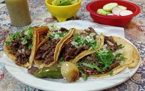 A plate of tacos filled with beef, onions, and cilantro, served with lime wedges and a side of cucumbers and radishes.