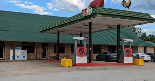 Gas station with two pumps, a green roof, and a stone building nearby. Ice sign visible on the left.
