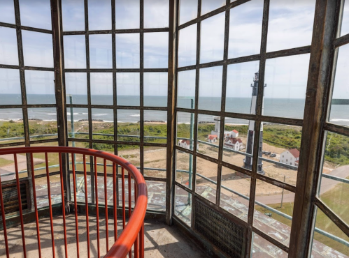 View from inside a lighthouse, featuring a spiral staircase and large windows overlooking the ocean and coastal landscape.