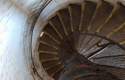 A spiral staircase with worn yellow steps, viewed from above, inside a cylindrical structure.