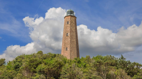 A tall lighthouse stands atop a green hill, surrounded by trees under a partly cloudy blue sky.