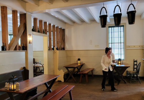 A cozy, rustic dining room with wooden tables, a woman serving, and lanterns on the tables. Buckets hang from the ceiling.