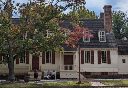 A historic house with white siding and red shutters, surrounded by trees, with two people sitting on the porch.