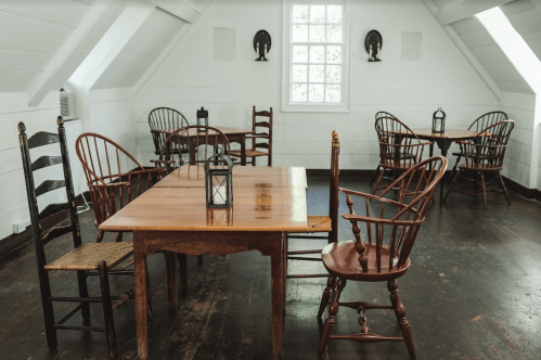 A cozy attic room with wooden tables and chairs, featuring lanterns and white walls.