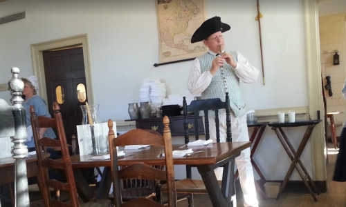 A man in historical attire plays a flute in a rustic dining room with wooden furniture and a map on the wall.