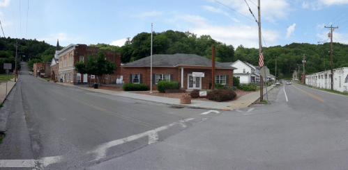 A street view of a small town with brick buildings, greenery, and a flagpole under a clear blue sky.