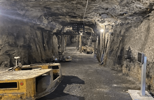 An underground mine tunnel with machinery and dim lighting, featuring rocky walls and a dirt floor.