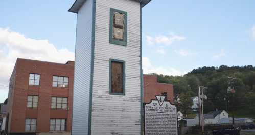A tall, weathered building with boarded windows next to a historical marker in a small town.