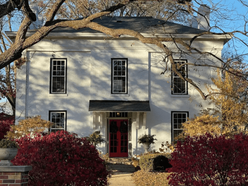 A white two-story house with a red door, surrounded by colorful autumn foliage and trees.