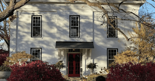 A white two-story house with a red door, surrounded by colorful autumn foliage and trees.