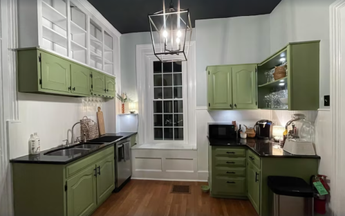 A modern kitchen with green cabinets, stainless steel appliances, and a large window, featuring warm lighting and wooden floors.