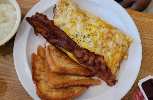 A plate with an omelette, crispy bacon, and toasted bread, served with a side of grits.
