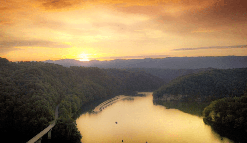 A serene sunset over a river, surrounded by lush green hills and mountains, reflecting golden hues on the water.