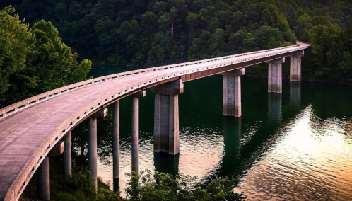 A curved concrete bridge spans over a calm lake, surrounded by lush green trees and reflecting the sunset.