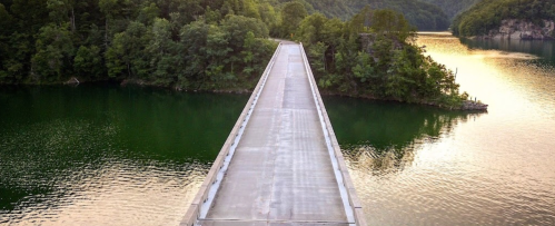 A long, straight road stretches over a calm lake, surrounded by lush green trees and reflecting golden light.